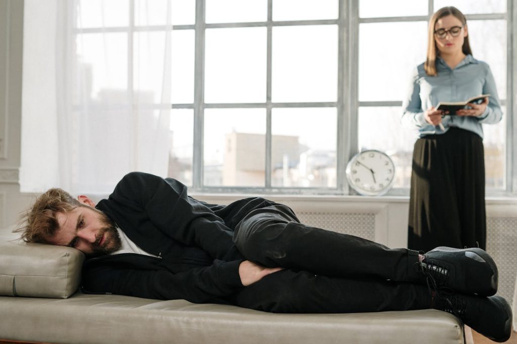 man-in-black-jacket-lying-on-bed-4101164 Psychological therapy session with a male patient lying on a couch and a female therapist taking notes.
