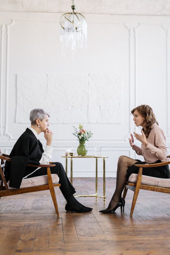 woman-in-black-long-sleeve-shirt-sitting-on-brown-wooden-chair-4098368 Two women engaged in a counseling session in a bright, modern loft setting.